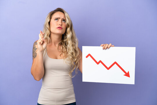 Young Brazilian woman isolated on purple background holding a sign with a decreasing statistics arrow symbol with fingers crossing