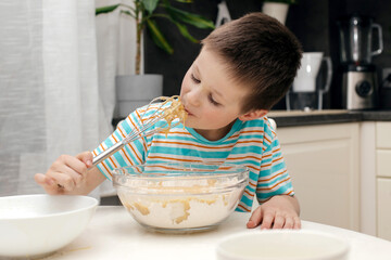 Cute boy licks a whisk with cake dough in the kitchen at home. Time with children at home. Little...