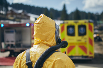 Member of biohazard team of emergency medical service in protective suit against ambulance and firefighters. - obrazy, fototapety, plakaty