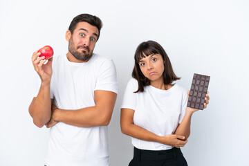 Young couple holding chocolate and apple isolated on white background making doubts gesture while lifting the shoulders