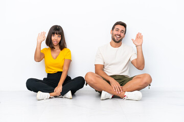 Fototapeta na wymiar Young couple sitting on the floor isolated on white background saluting with hand with happy expression