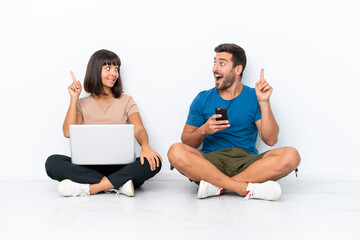 Obraz premium Young couple sitting on the floor holding pc and mobile phone isolated on white background intending to realizes the solution while lifting a finger up