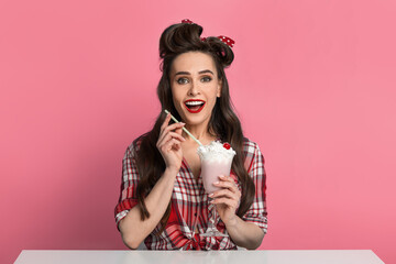 Excited young pinup woman in retro style clothes drinking milk shake from glass, sitting at table over blue background