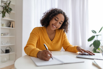Young black woman studying or working online with laptop at home, taking notes, enjoying distance job or education