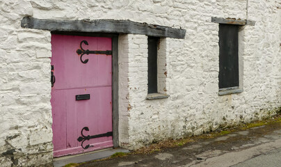 Abandoned, boarded up frontage of an old cottage. Original weathered oak lintels above windows and door. Pink painted stabledoor entrance with old ironwork furniture. Whitewashed exterior with moss. - 502026973