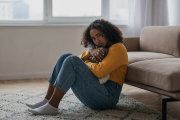 Young black woman with depression sitting on floor and hugging pillow at home