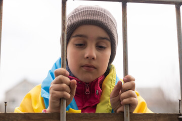 sad little girl with the flag of Ukraine behind a metal fence. Social problem of refugees and...