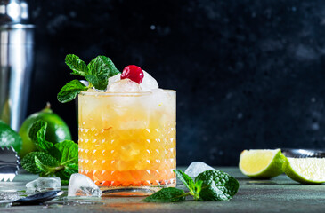Mai Tai popular alcoholic cocktail with rum, liqueur, syrup, lime juice, mint and crushed ice. Dark green background, steel bar tools, negative space