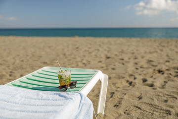 A glass of chilled cocktail mohito and sunglasses on deck chair on a beach
