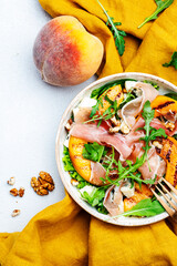 Summer salad bowl with sweet grilled peach, jamon, soft cheese, walnuts and fresh arugula on white kitchen table background, top view, negative space