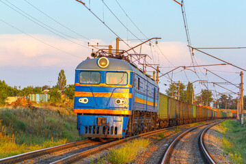 A blue locomotive with a long freight train is going to the railway station. Freight railway transportation of metal products. Sunset lighting.