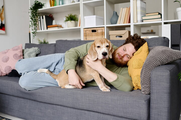 Young bearded man lying on sofa and embracing his dog during leisure time at home - obrazy, fototapety, plakaty