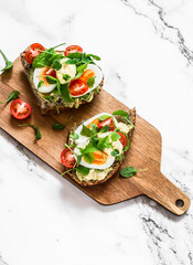 Vegetarian sandwiches with grain bread, avocado, boiled egg, microgreens, cherry tomatoes and homemade mayonnaise on a cutting board on a light background