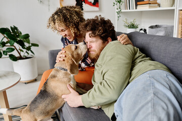 Cute dog licking face of owner while he lying on sofa with girl laughing in background