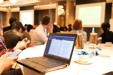 desktop computer, mobile phone, glass, water bottle on a table with blurred meeting participants and screen in background in a conference hall.