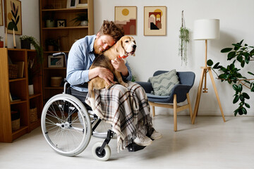 Fototapeta na wymiar Young bearded man embracing with his lovely dog while sitting in wheelchair in living room