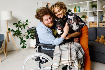 Fototapeta na wymiar Young bearded man in wheelchair spending rehabilitation at home together with his wife and lovely dog