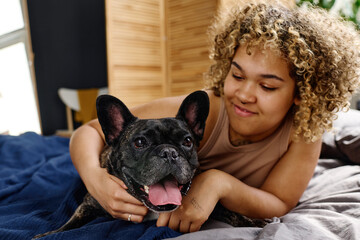 African young girl with curly hair embracing her french bulldog during rest time in bed