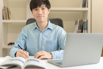 Young Asian college male student writing notes, using laptop for learning online education, business learning, watching online webinar. Businessman is trading, investing on online platform on laptop.