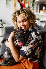 African young girl with curly hair sitting on sofa holding her dog on arms, she bonding with her pet
