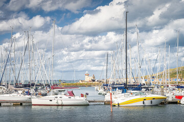 Naklejka premium Howth lighthouse seen through masts of sailboats, yachts and motorboats moored in Howth marina, Dublin, Ireland