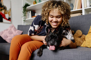 African young woman with curly hair happy to have a dog, she sitting on sofa and caressing her...
