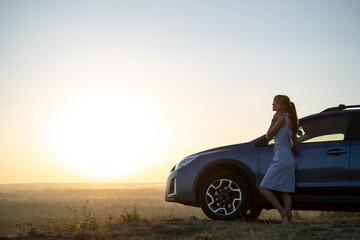 Happy woman driver in blue summer dress enjoying warm evening near her car. Travel and vacations concept