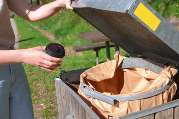 A woman throws a cardboard cup after coffee with a plastic lid into a paper container.