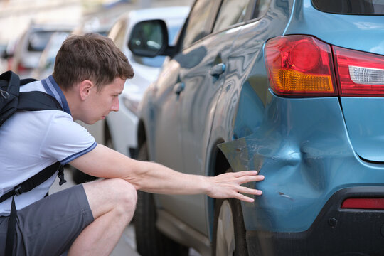 Sad young man driver sitting near his dented car looking shocked on damaged fender in road accident