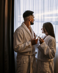 Couple wearing bathrobes in hotel standing near window with glasses of champagne