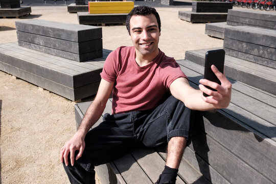 Smiling young man sitting in modern bench taking a selfie with mobile.