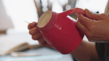 Hands of an elderly woman painting clay mug in red color