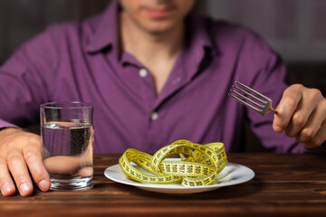 Diet concept. Measuring tape on plate. Man eating measuring tape with fork with glass of water