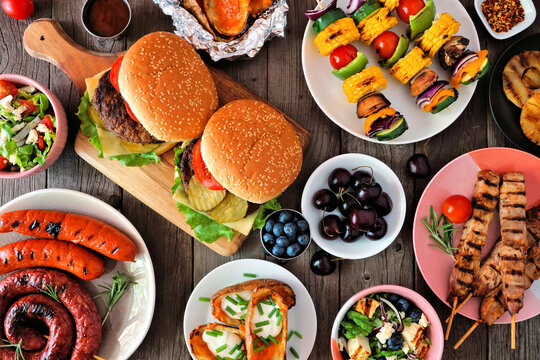 Summer BBQ or picnic food table scene. Selection of burgers, grilled meat, vegetables, fruits, salad and potatoes. Above view on a dark wood background.