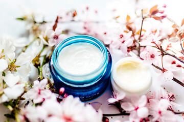 Obraz na płótnie Canvas Top view of cosmetic cream and concealer eyes creams with pink cherry flowers in a blue glass jar. Hygienic skincare lotion product. Horizontal photo