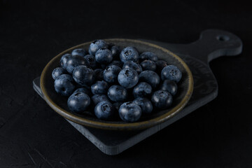 Fresh organic blueberries in a ceramic plate. The plate stands on a gray board on a black background. Close-up.