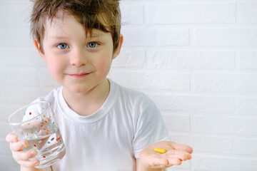 the boy is looking at the camera, holding a glass of water and a yellow pill in his hand. The child...