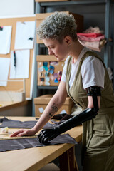 Young businesswoman with disability looking at piece of textile while standing by table and working...