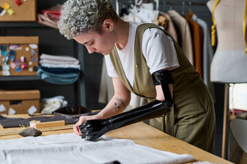 Young female tailor bending over table with piece of fabric while working over order of client in large workshop or studio of fashion design