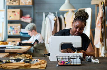Young African American businesswoman using electric sewing machine while sitting by workplace...