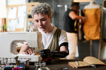Young serious seamstress with partial arm sewing new clothes on electric machine while working over...