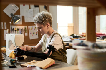 Young serious female tailor with partial arm concentrating on sewing work while sitting by table in front of electric machine