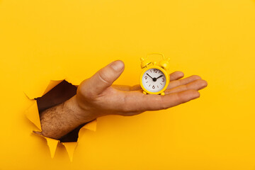 Hand with mini alarm clock through a paper hole in yellow background