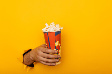 Male hand holds popcorn through torn hole yellow paper with copy space. Entertainment concept