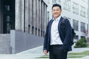 Successful asian business tourist looking at camera and smiling, man in business suit and backpack