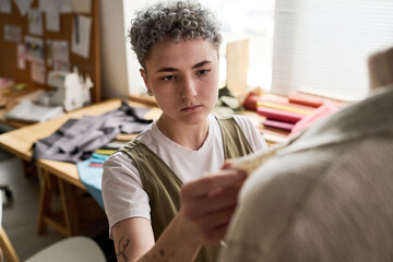 Young serious seamstress with grey short curly hair standing in front of mannequin while trying on unfinished attire in workshop