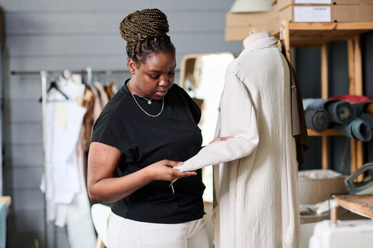 Young Contemporary Black Woman In Casualwear Checking Sewing Quality Of White Coat On Mannequin While Looking At Sleeve In Workshop