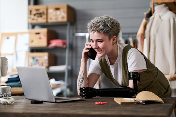 Happy young female tailor speaking on mobile phone with client while looking through new orders on laptop screen by workplace - obrazy, fototapety, plakaty