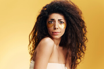 Confused Funny tanned pretty Latin lady in gold hydrogel patches under eye posing isolated over yellow background. Cosmetic product ad Natural beauty concept Studio portrait Copy space