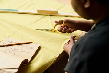 Hand of young black woman with scissors cutting thick yellow textile by workplace while creating...